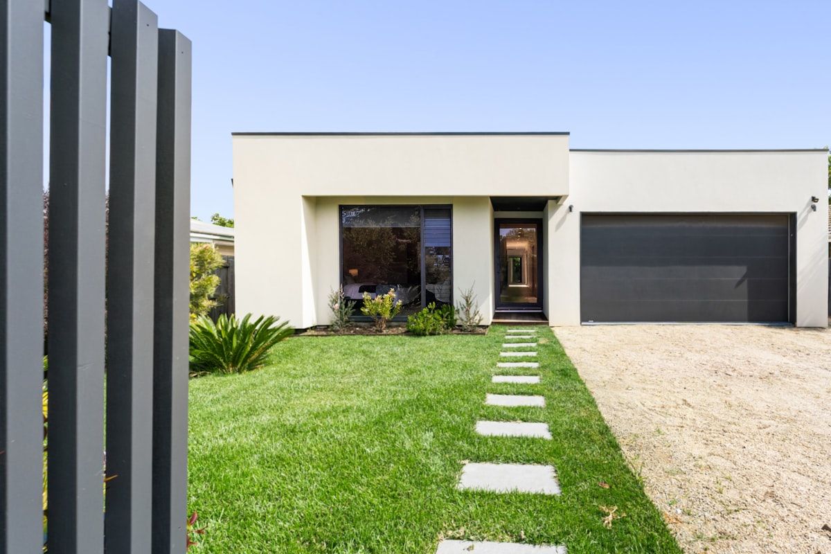 A modern front property with three gate panels displayed in a neat row: a light aluminium slat gate, a darker steel gate with a powder-coated finish, and a timber-look gate with horizontal boards. Behind them is a contemporary house facade with matching fence sections and brick piers, suggesting how the gate design coordinates with the home and streetscape.