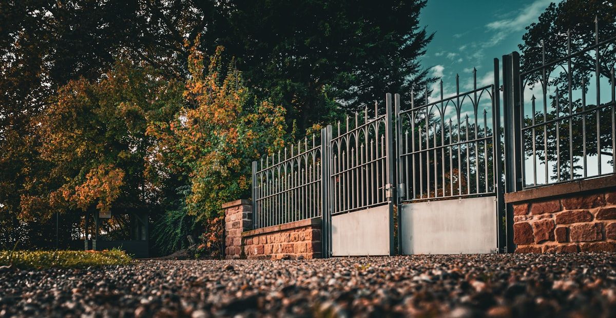 A row of driveway gates showing different finishes: a light aluminium slat gate, a darker steel gate with a solid frame, and a timber-look aluminium gate in front of homes with mixed brick and modern facades, with one gate set near a coastal-style setting and another beside a tidy inland front yard.