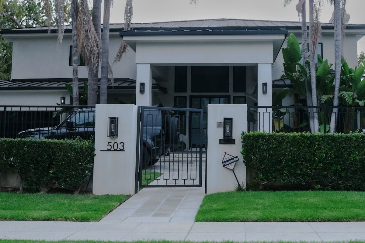 A close-up view of an automated driveway gate installation showing a gate motor mounted at the base of a sliding gate, a keypad on a fence post, an intercom unit near the entrance, and safety sensors positioned low beside the opening on a residential driveway.