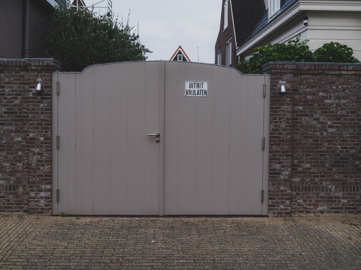 A partially completed driveway gate project at a suburban home, with steel gate panels laid on sawhorses, concrete footings exposed beside brick piers, trenching marks for electrical work, a powder-coated gate section leaning against fencing, and installed hardware arranged nearby for final assembly.