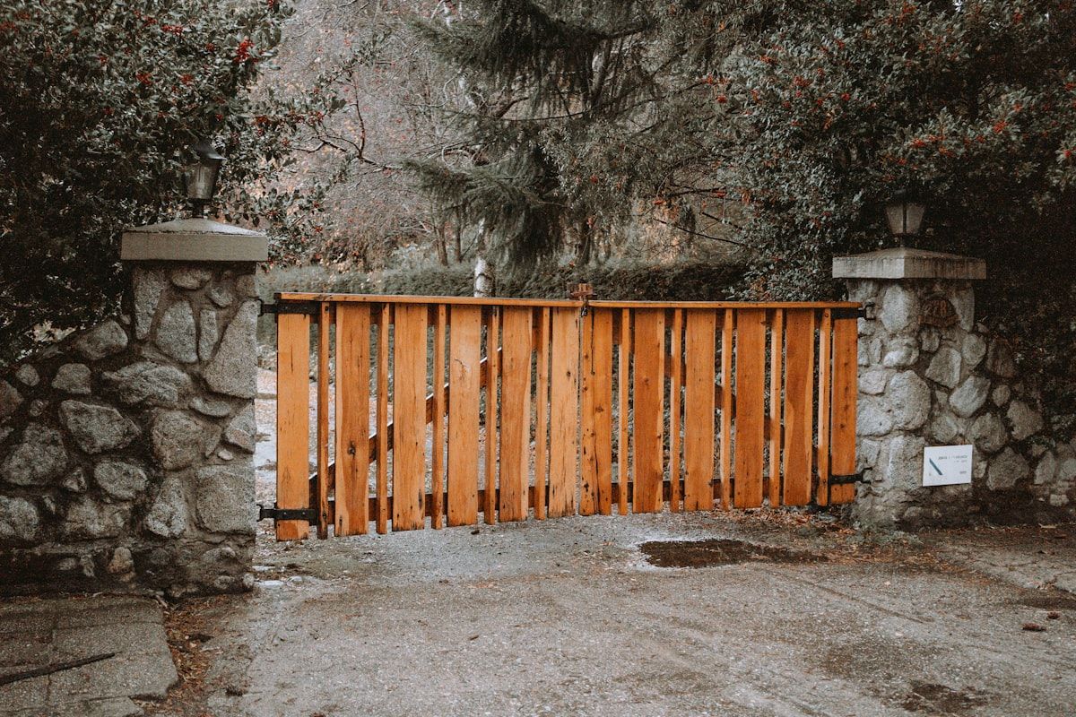 A residential gate installation site under construction with a partially installed sliding gate, concrete footings, exposed electrical conduit in a trench, stacked fence materials, and a crew preparing posts near a wide driveway entrance. The ground shows excavation marks and drainage work, visually representing the separate elements that contribute to the final cost.