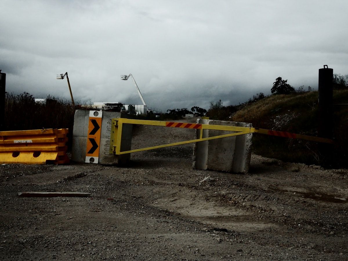 A close view of a gate installation job in progress with stacked aluminium gate panels, a steel gate frame, concrete footings being poured near new posts, conduit trenches beside the driveway, and a technician positioning automation hardware at the opening