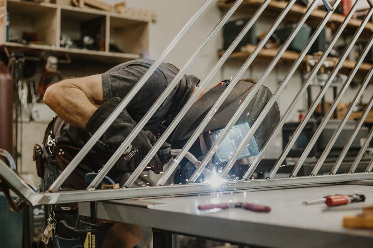 A workshop-style scene with a partially assembled metal gate frame on stands, a heavy-duty gate motor, rollers, hinge hardware, electrical cable, concrete posts, and a worker’s measuring tools laid out beside the materials for an installation quote discussion.